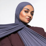 A woman wearing a slate blue-gray hijab and a matching dark plum-brown top, photographed against a clean light gray studio background.
