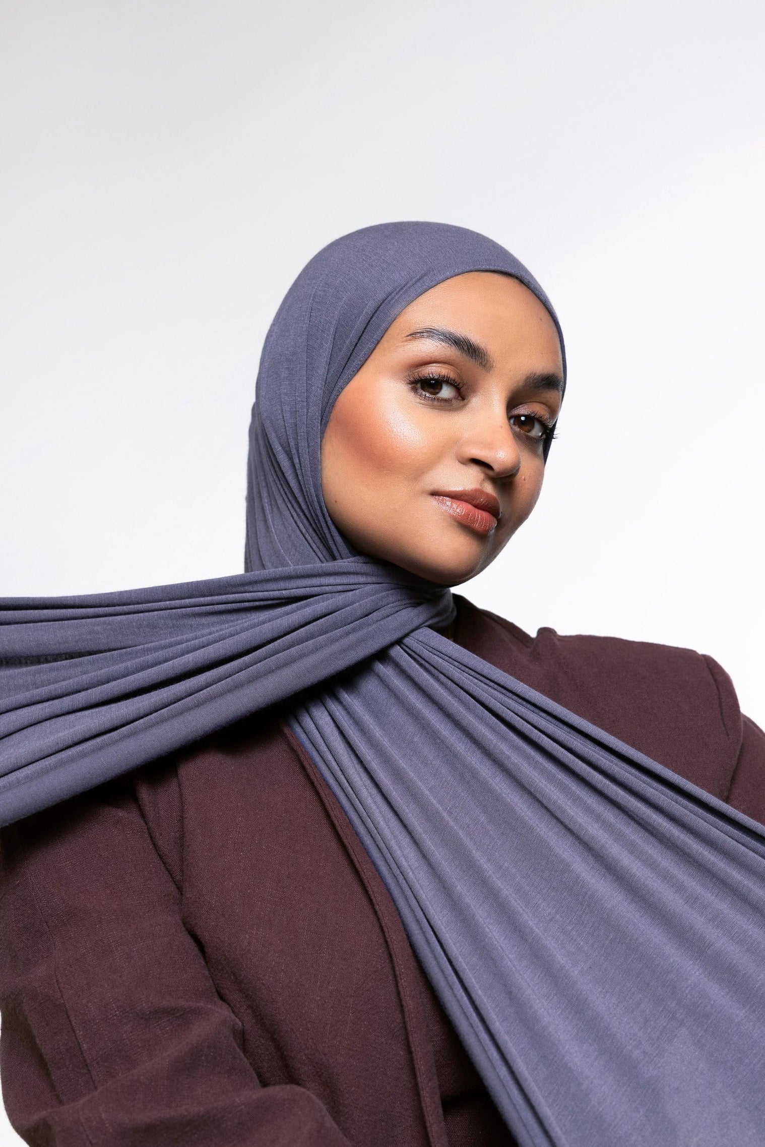A woman wearing a slate blue-gray hijab and a matching dark plum-brown top, photographed against a clean light gray studio background.