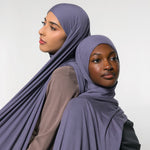 Two women wearing draped mauve-purple hijabs, posed against a plain light gray studio background.