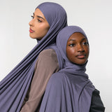 Two women wearing draped mauve-purple hijabs, posed against a plain light gray studio background.