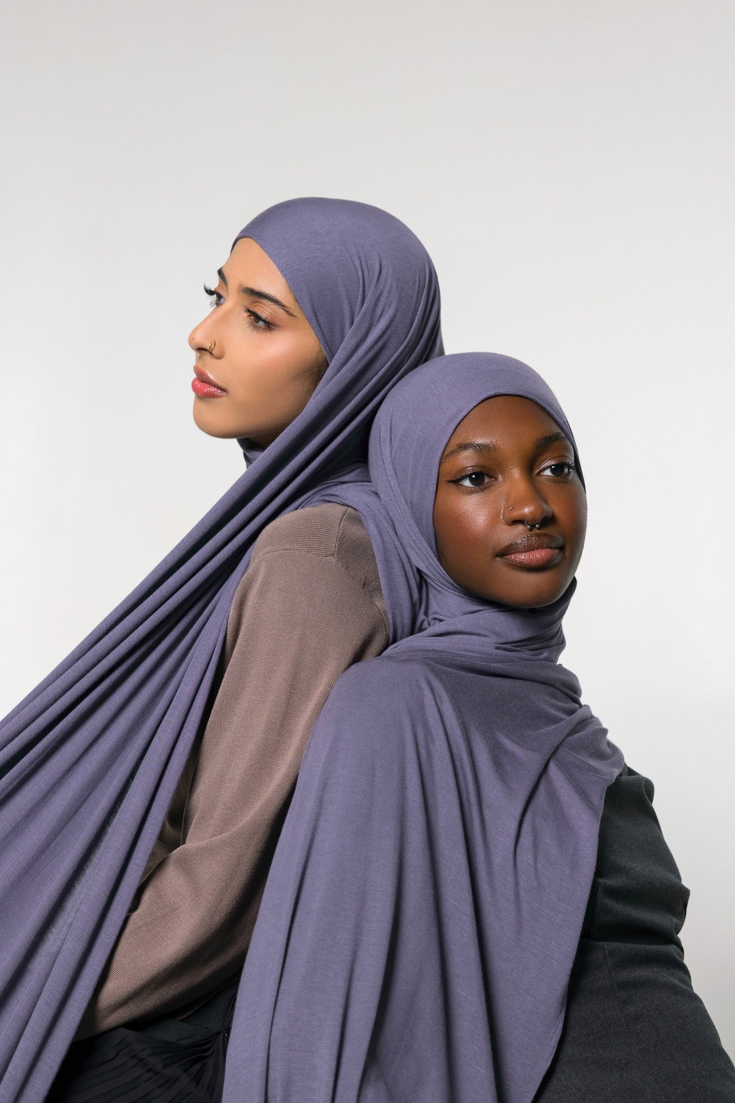 Two women wearing draped mauve-purple hijabs, posed against a plain light gray studio background.