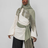 A model wearing a light olive green hijab and matching fringed wrap with a tie belt, styled over a cream long-sleeve top and black wide-leg pants, against a light gray studio background.