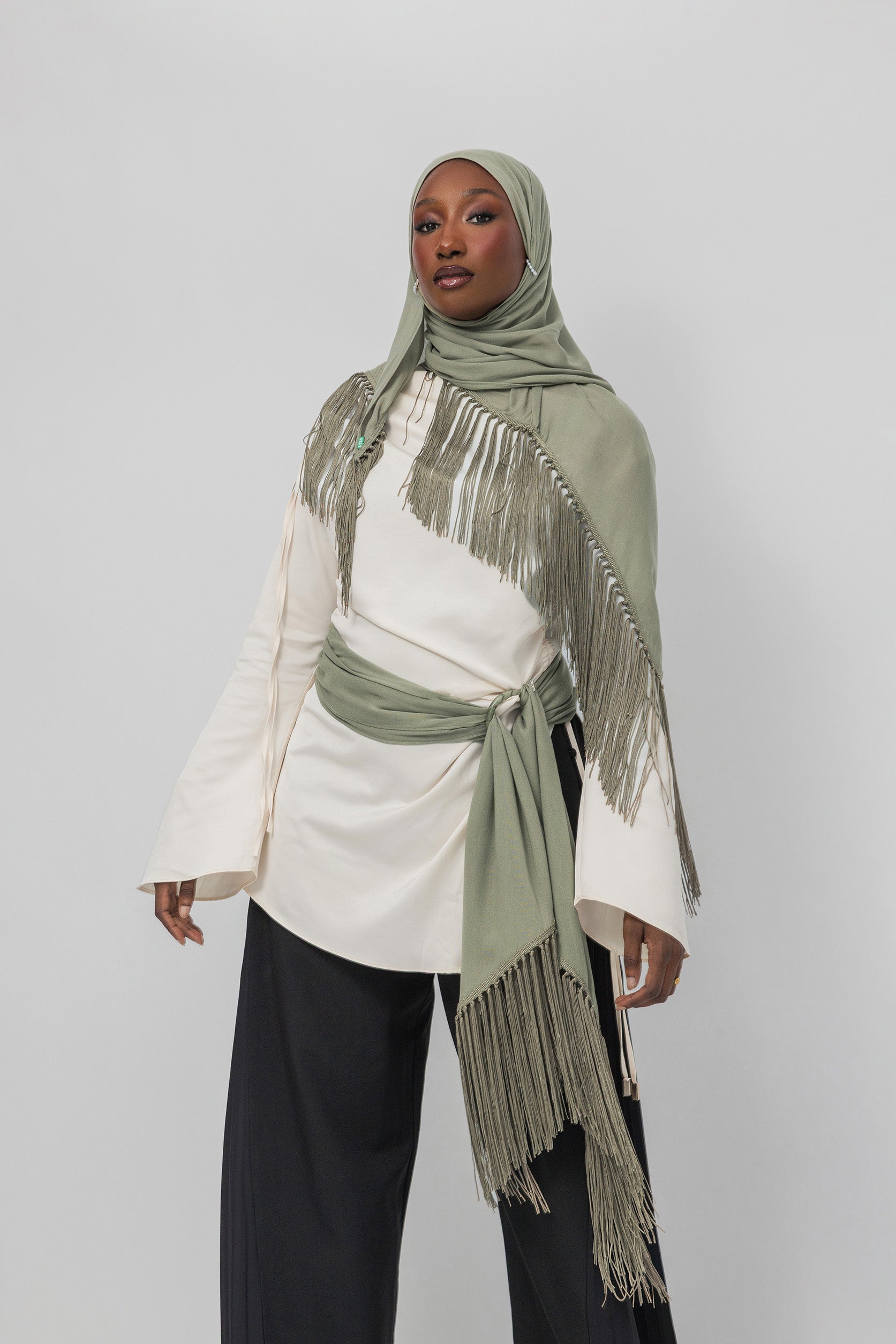 A model wearing a light olive green hijab and matching fringed wrap with a tie belt, styled over a cream long-sleeve top and black wide-leg pants, against a light gray studio background.