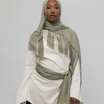 A woman wearing a light olive green fringed hijab and a matching tied sash over an off-white long-sleeve top, paired with black pants, against a plain light gray studio background.