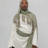 A woman wearing a light olive green fringed hijab and a matching tied sash over an off-white long-sleeve top, paired with black pants, against a plain light gray studio background.