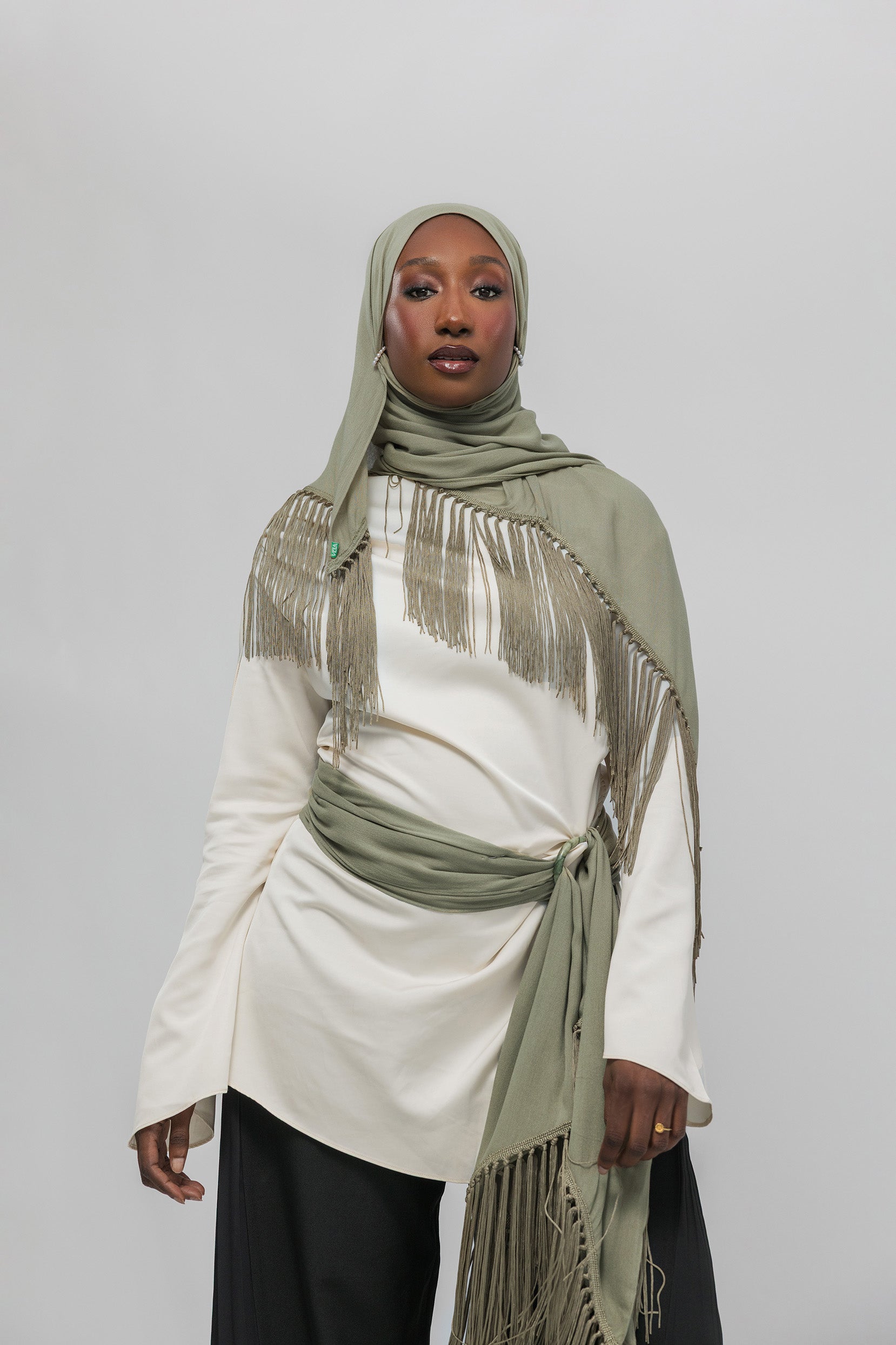 A woman wearing a light olive green fringed hijab and a matching tied sash over an off-white long-sleeve top, paired with black pants, against a plain light gray studio background.