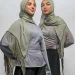 Two women modeling matching light olive green hijabs and shawls with fringe details over gray tops and black pants, photographed against a plain light gray studio background.