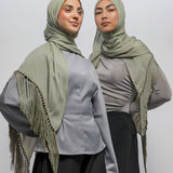 Two women modeling matching light olive green hijabs and shawls with fringe details over gray tops and black pants, photographed against a plain light gray studio background.