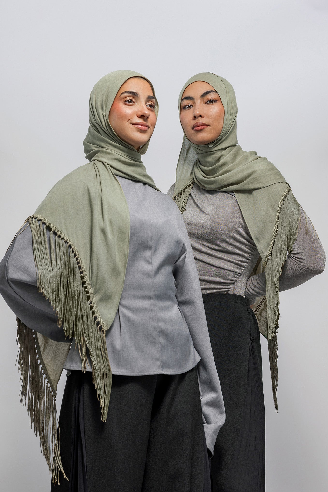 Two women modeling matching light olive green hijabs and shawls with fringe details over gray tops and black pants, photographed against a plain light gray studio background.
