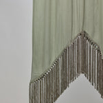 A light olive green draped fabric accessory with a pointed triangular tip and long fringe trim along the edge, shown against a plain light gray background.