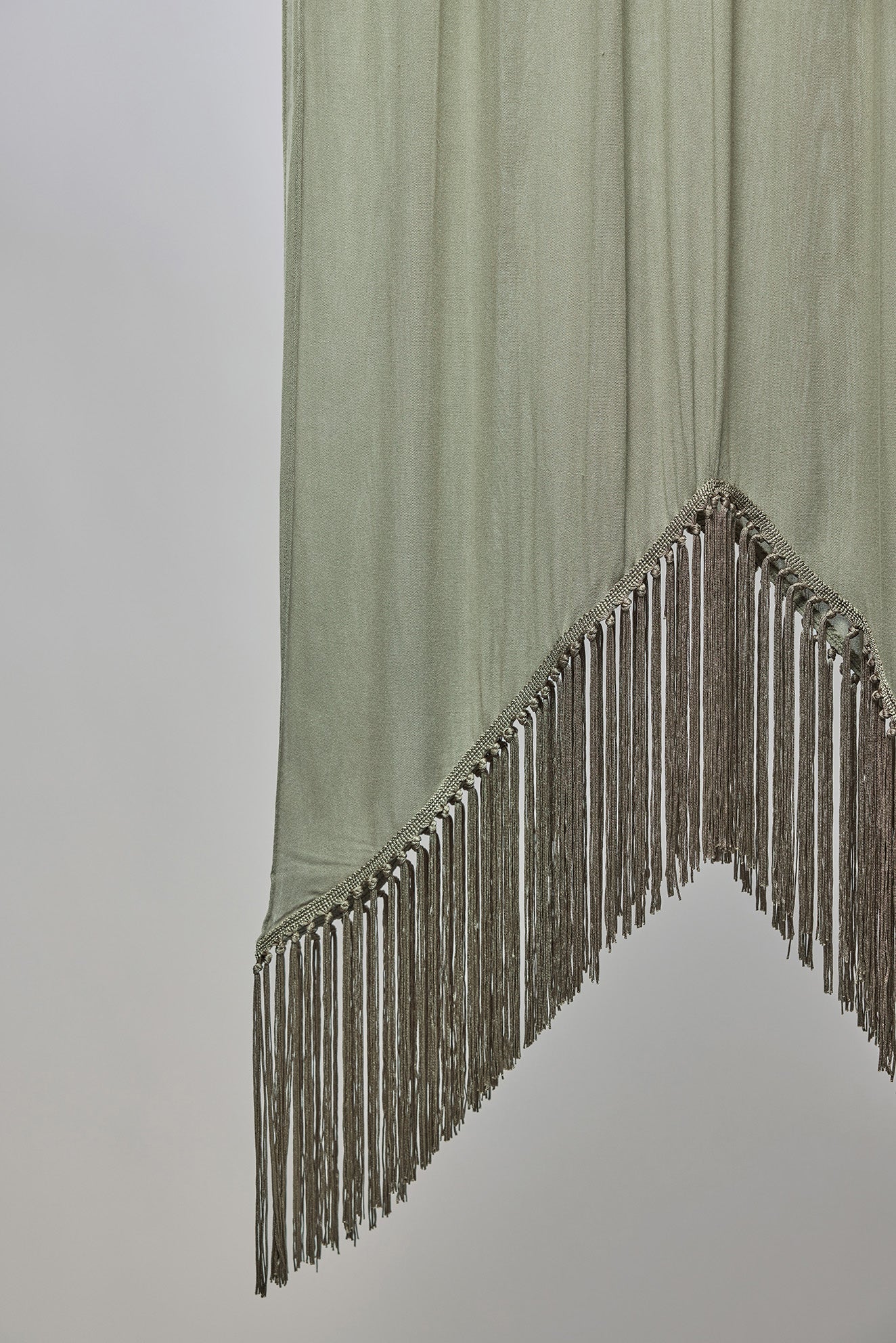 A light olive green draped fabric accessory with a pointed triangular tip and long fringe trim along the edge, shown against a plain light gray background.