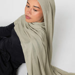 A woman wearing a light sage green hijab and a black long-sleeve top, posed against a plain light gray background.