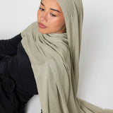 A woman wearing a light sage green hijab and a black long-sleeve top, posed against a plain light gray background.