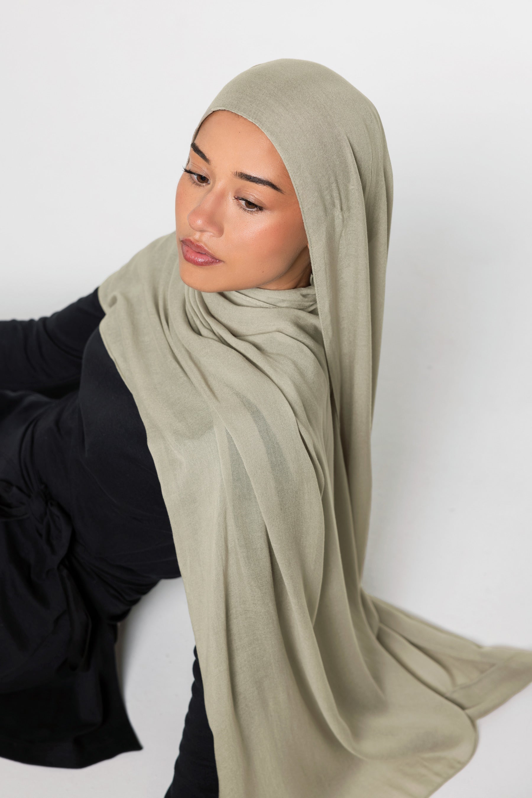 A woman wearing a light sage green hijab and a black long-sleeve top, posed against a plain light gray background.
