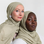 Two women wearing matching sage green hijabs, posed against a clean white studio background.