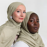Two women wearing matching sage green hijabs, posed against a clean white studio background.