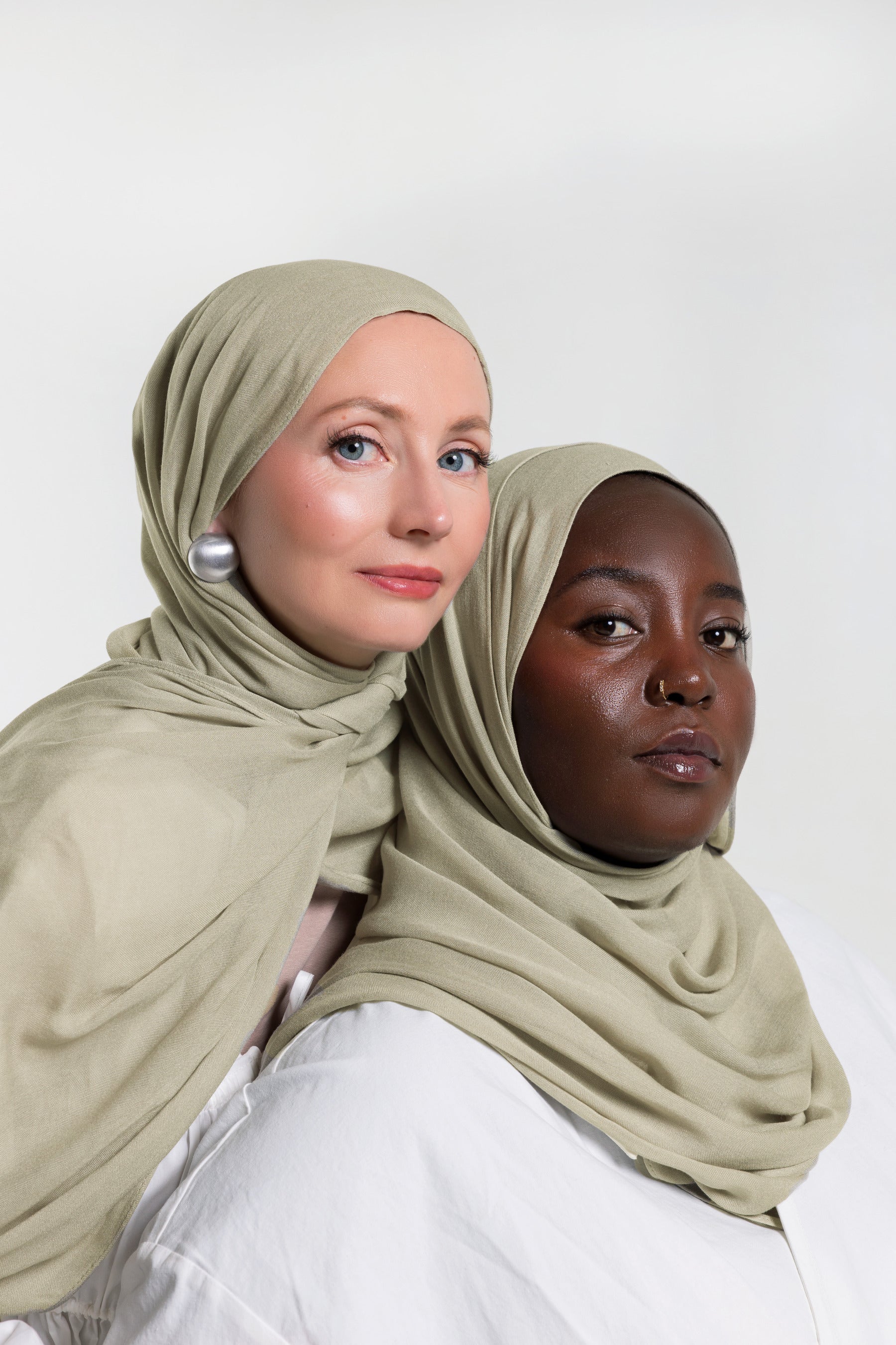Two women wearing matching sage green hijabs, posed against a clean white studio background.
