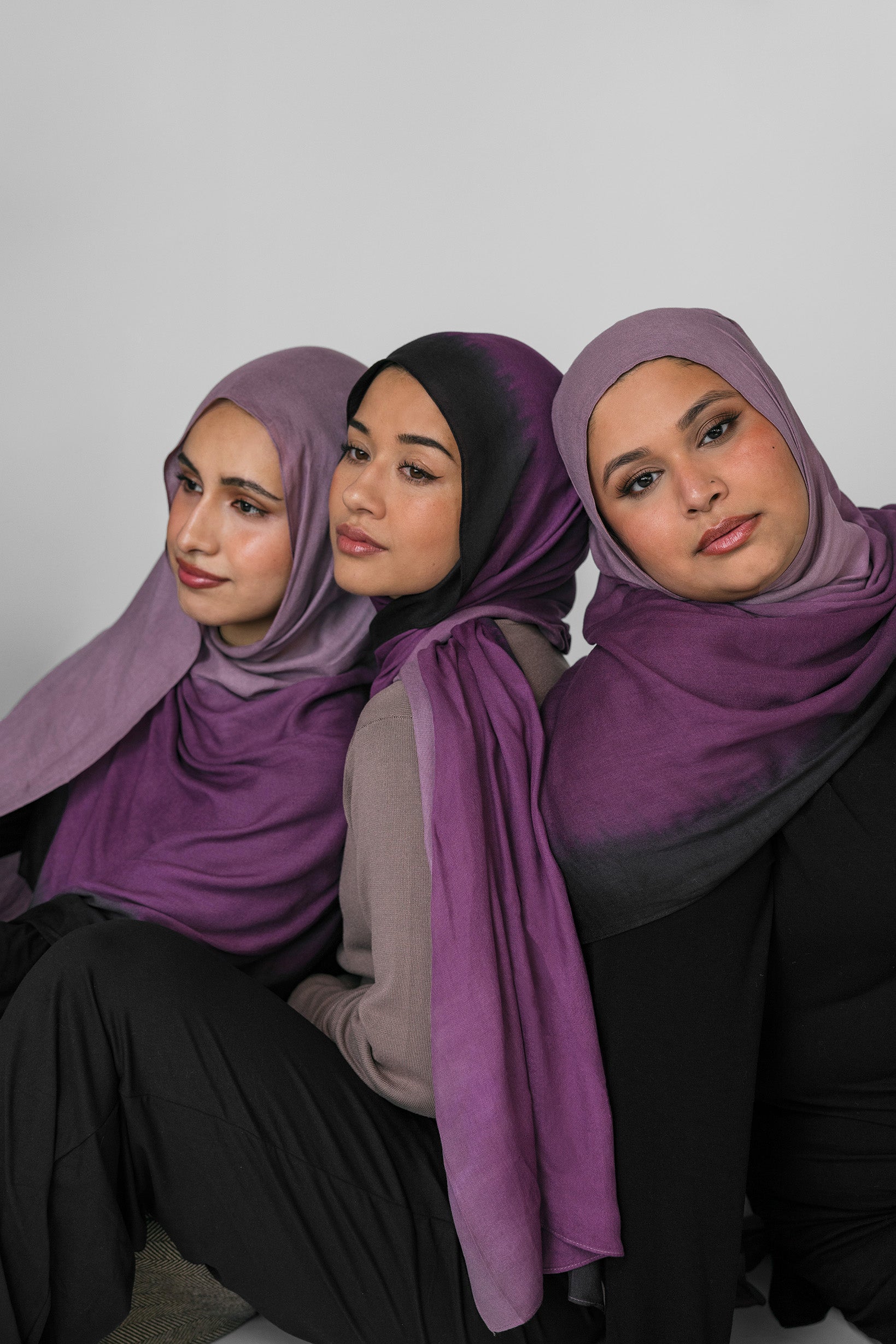 Three women wearing purple ombre hijabs sitting side by side against a plain background