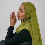 Woman wearing a green hijab with gold embellishments against a plain background