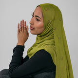 Woman wearing a green hijab with gold embellishments against a plain background