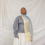 Person wearing a gray top, white skirt, and colorful blue / yellow hijab against a plain background