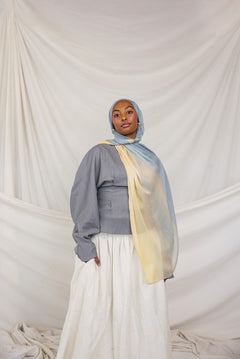 Person wearing a gray top, white skirt, and colorful blue / yellow hijab against a plain background