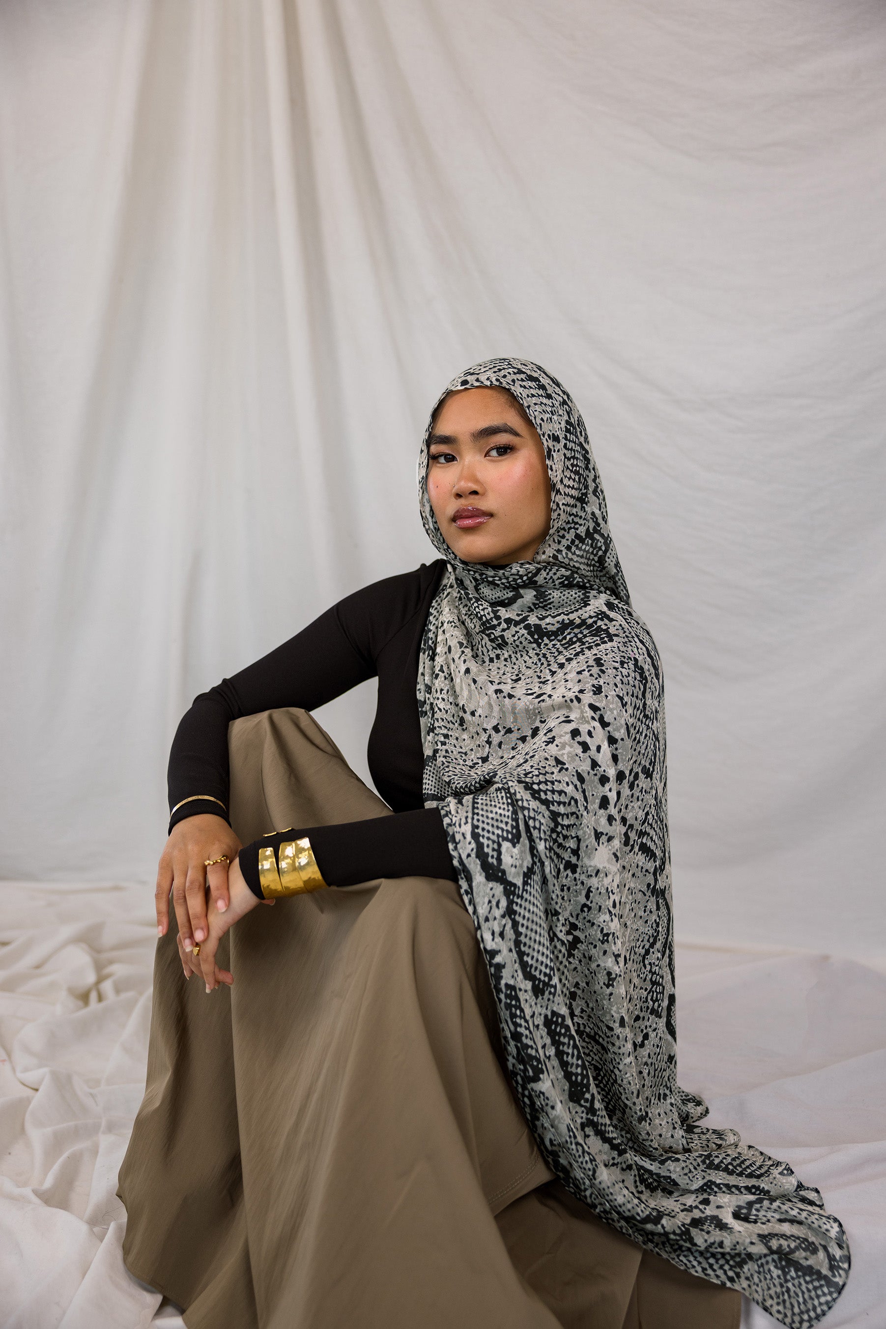 Model shown sitting down and posing in Ink Cobra, a grey-toned snakeskin print scarf by VELA. 