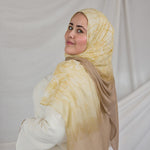 Woman wearing a yellow beige shell printed hijab and white outfit against a plain background