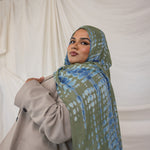 Woman wearing a green and blue ripple patterned hijab and beige coat against a white curtain background