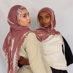 Two women wearing pink rippled printed hijabs and neutral-colored clothing against a plain background