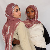 Two women wearing pink rippled printed hijabs and neutral-colored clothing against a plain background