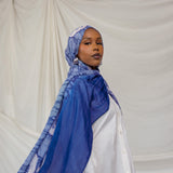 Woman wearing a royal blue and white shell printed headscarf against a plain background
