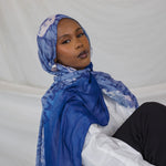 Model wearing a blue hijab with white shell prints and sitting on a white surface
