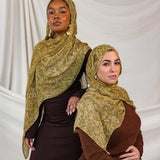 Models shown posing in the Golden Cobra scarf by VELA. This image features one model standing and one sitting, both wearing the golden/yellow toned snakeskin print. 