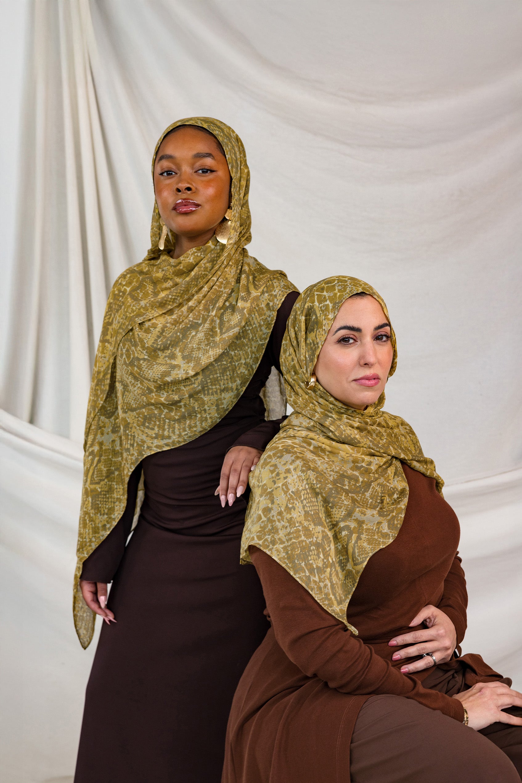 Models shown posing in the Golden Cobra scarf by VELA. This image features one model standing and one sitting, both wearing the golden/yellow toned snakeskin print. 