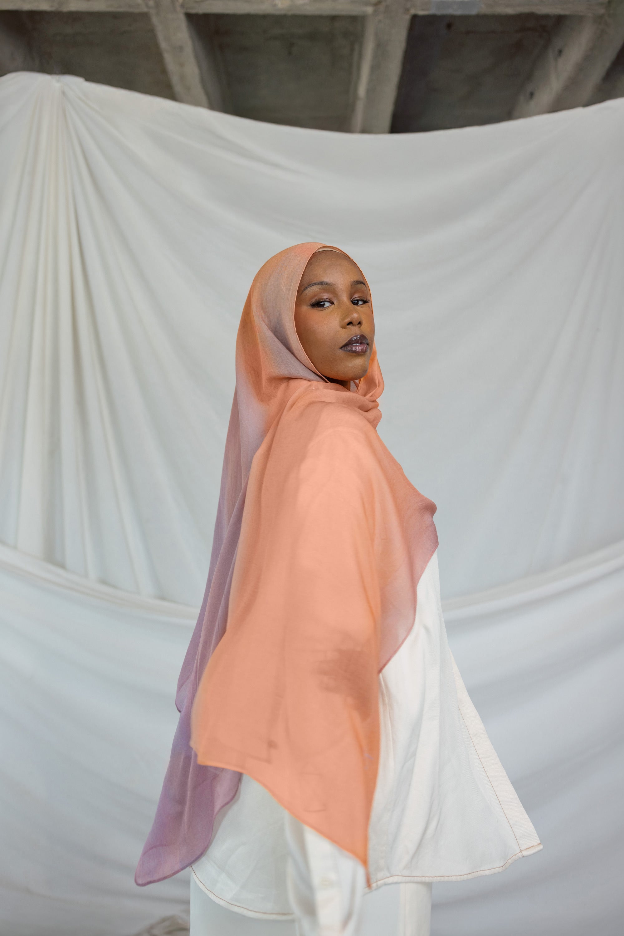 Woman wearing a bright pink / purple pastel hijab in front of draped fabric