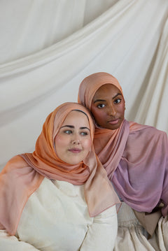 Two women wearing pink and purple ombre hijabs against a white curtain background