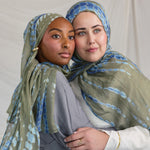 Two women wearing green and blue patterned headscarves embracing each other against a plain background