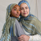 Two women wearing green and blue patterned headscarves embracing each other against a plain background