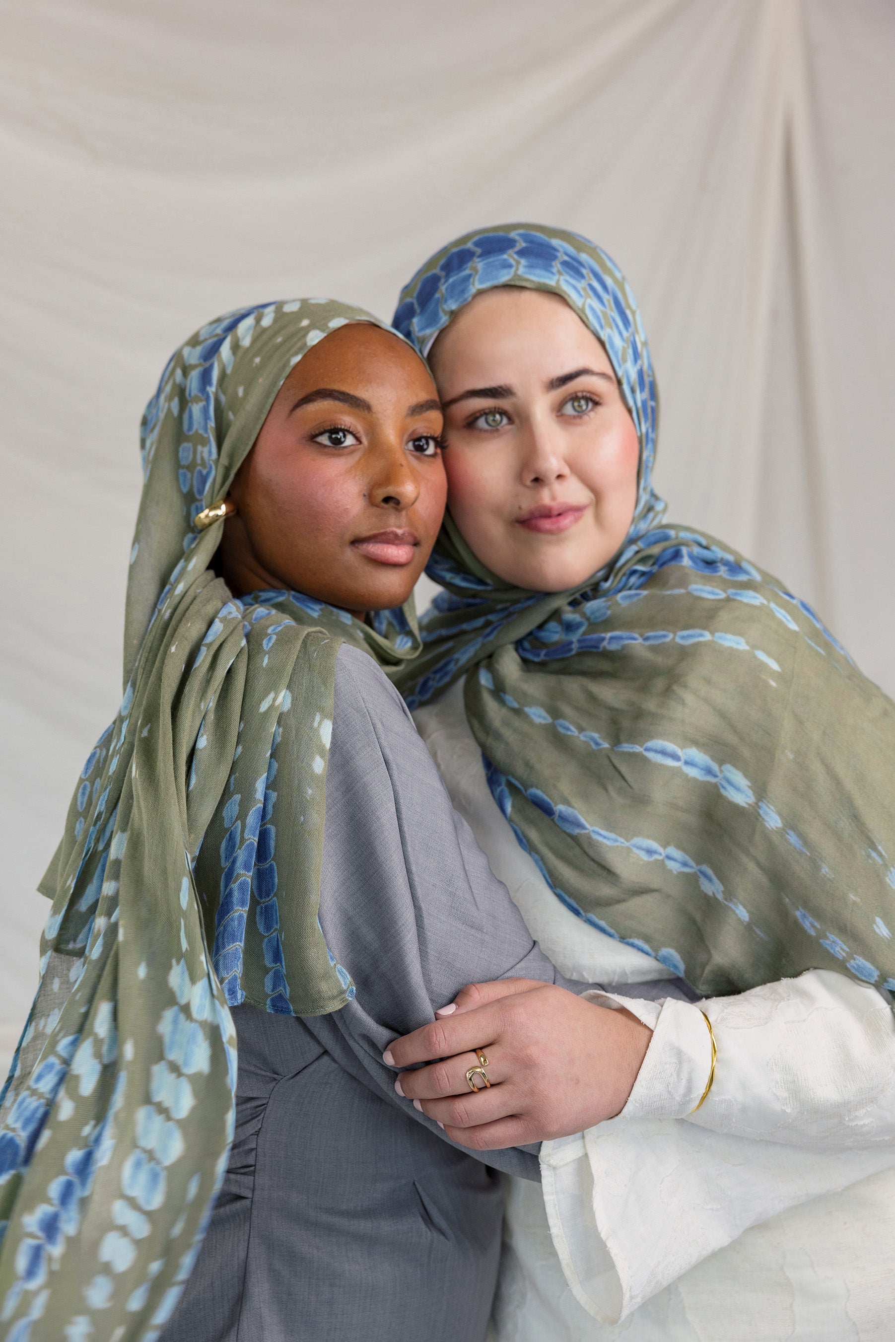Two women wearing green and blue patterned headscarves embracing each other against a plain background