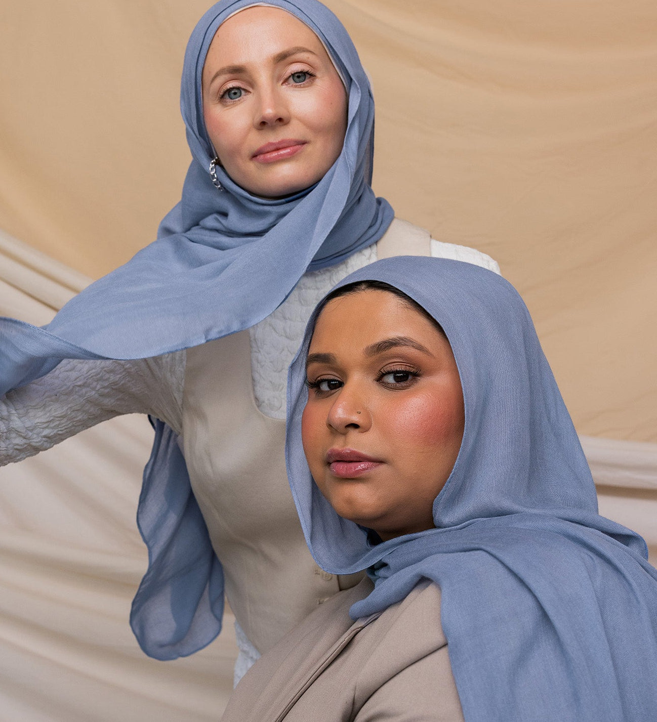 Two women wearing blue hijabs with gray undertones, standing in front of a neutral backdrop.