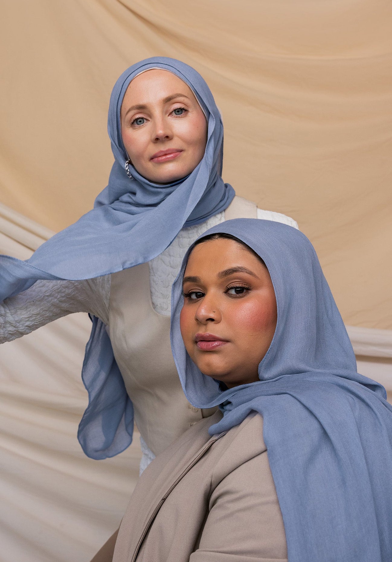 Two women wearing blue hijabs with gray undertones, standing in front of a neutral backdrop.