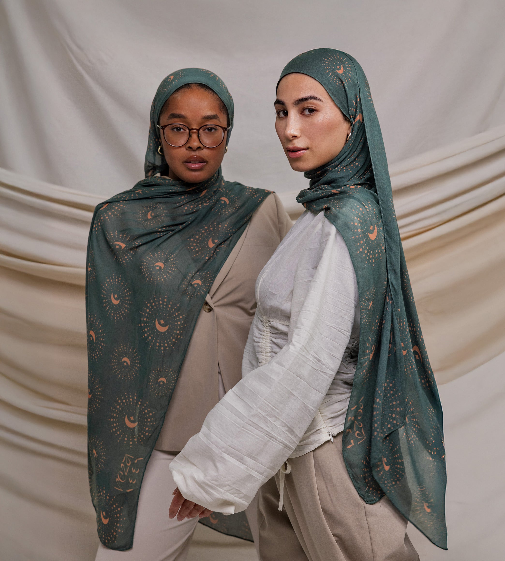 Two models wearing a teal woven hijab with crescents on it. the hijab is draped on the models around their head. the models are standing in front of a cream fabric backdrop.