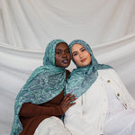 Two models shown wearing the Aqua Cobra scarf by VELA, featuring a snakeskin print in teal and blue. 