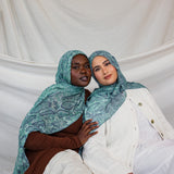 Two models shown wearing the Aqua Cobra scarf by VELA, featuring a snakeskin print in teal and blue. 