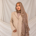 Woman wearing a beige coat and mocha patterned hijab against a plain background
