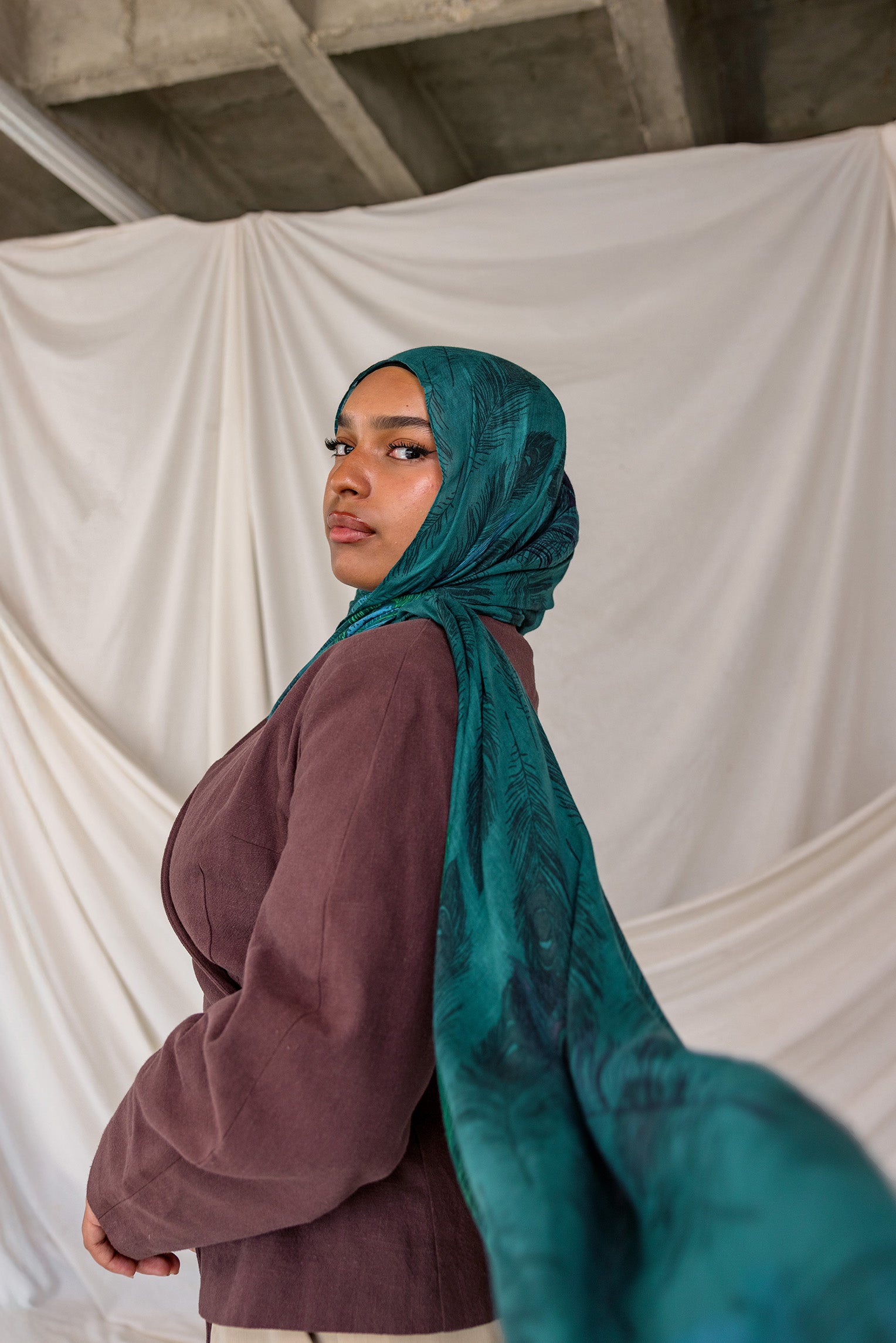Model wearing a teal peacock inspired printed hijab and brown top against a white backdrop