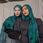 Two women wearing peacock feather inspired headscarves and dark clothing against a plain background