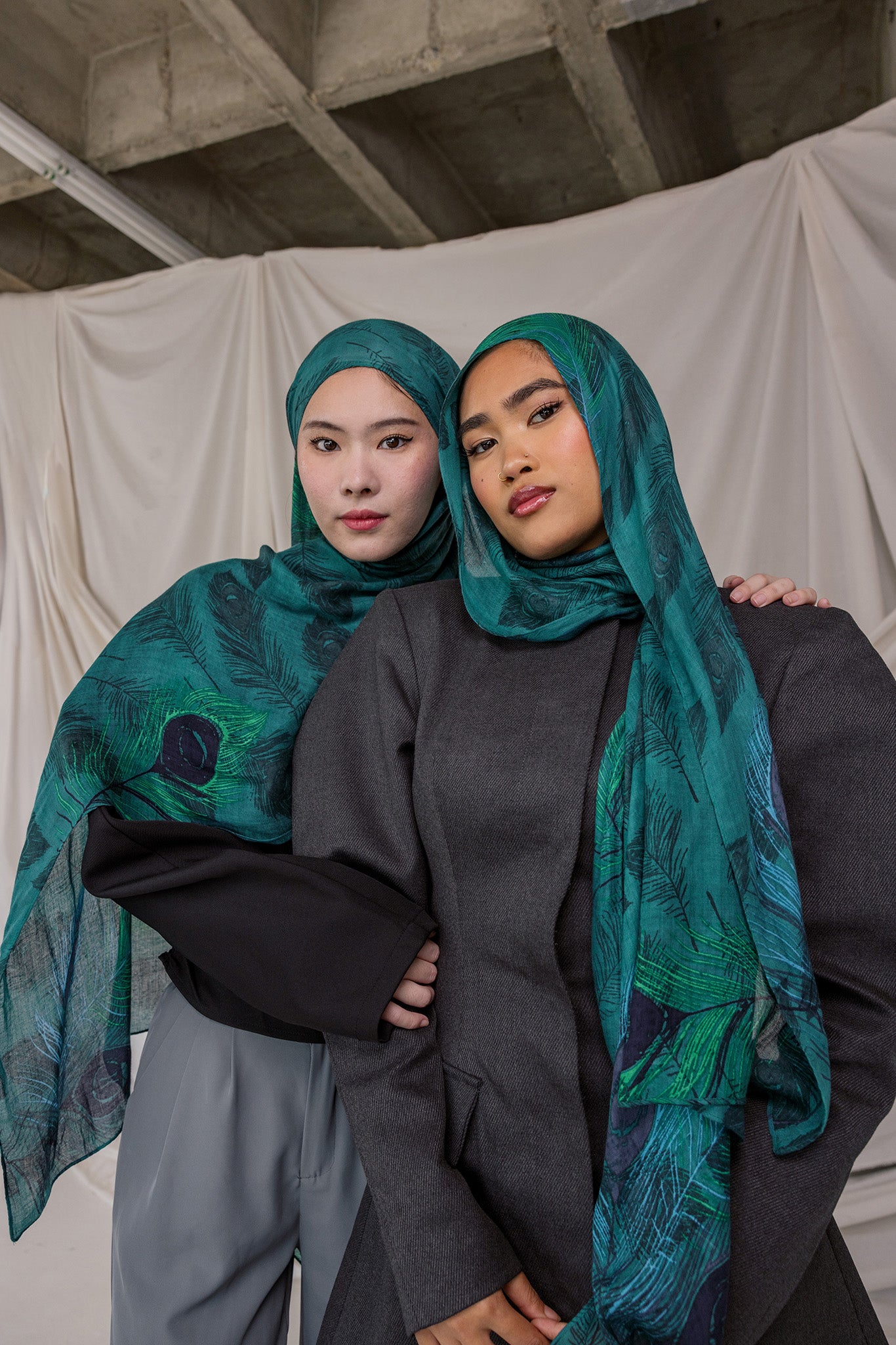 Two women wearing peacock feather inspired headscarves and dark clothing against a plain background