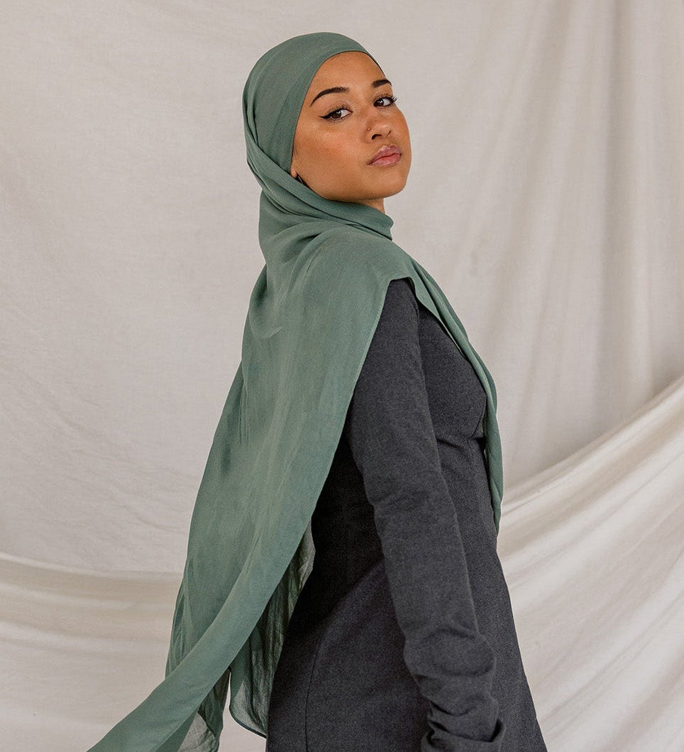 Person wearing a green hijab, dark top, and gray pants against a beige draped curtain background